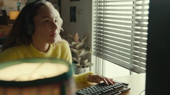一个身穿黄色毛衣、头戴耳机的人正用键盘打字。前景中有盏台灯。